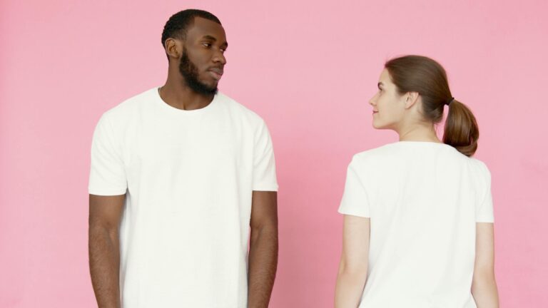 A man and woman in white t-shirts stand against a pink backdrop.